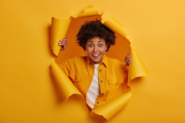 Joyeuse femme ethnique se sent heureuse, se tient à travers le trou déchiré de fond jaune