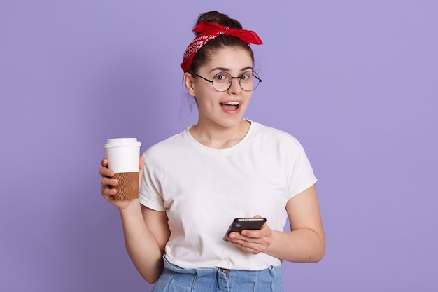 Joyeuse femme européenne joyeuse en t-shirt décontracté blanc et bandeau rouge, tenant le café à emporter et la navigation sur Internet dans le téléphone mobile, SMS ami,