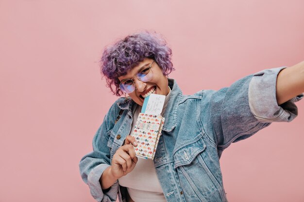 Joyeuse femme lumineuse aux cheveux courts bouclés lilas dans des verres ronds détient des billets et prend selfie sur rose isolé.