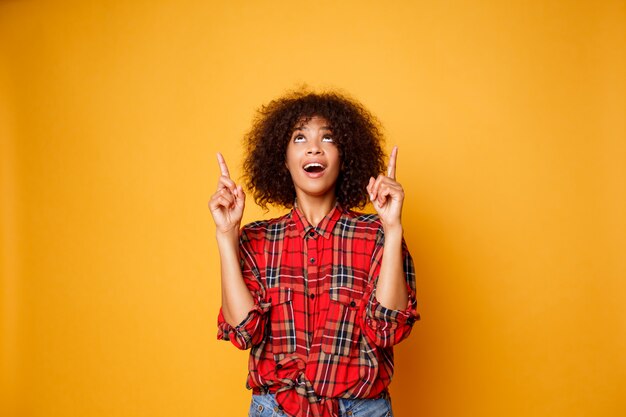 Joyeuse femme noire américaine en chemise rouge regardant vers le haut et pointant les doigts vers le haut sur l'espace de copie isolé sur fond orange.