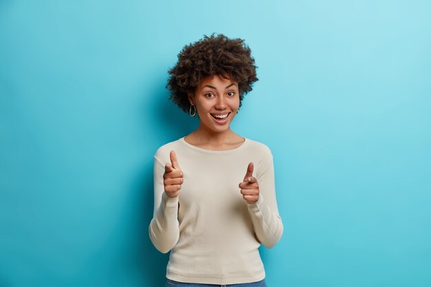 Joyeuse femme à la peau sombre avec des cheveux bouclés sélectionne quelqu'un fait un geste de pistolet à doigt