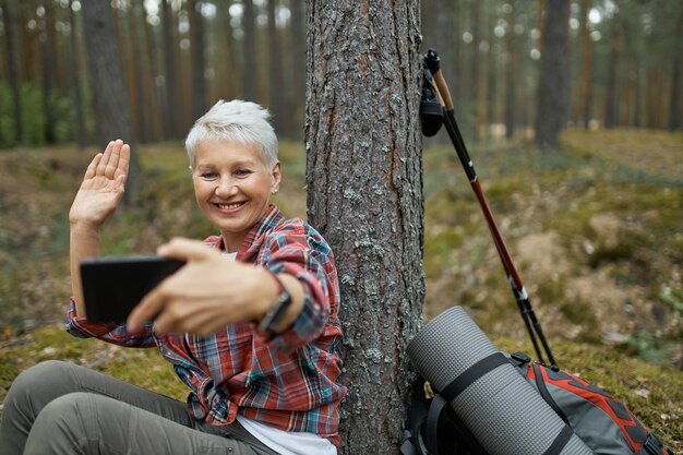 Joyeuse femme retraitée active assise sous un arbre avec du matériel de randonnée tenant un téléphone portable, souriant et agitant la main, parlant à son amie par vidéoconférence à l'aide de l'application en ligne.