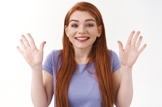 Joyeuse femme rousse souriante sortante en t-shirt, levant les mains et les agitant en bonjour, salut geste, saluant joyeusement un ami, voir une personne familière, dire au revoir, mur blanc debout