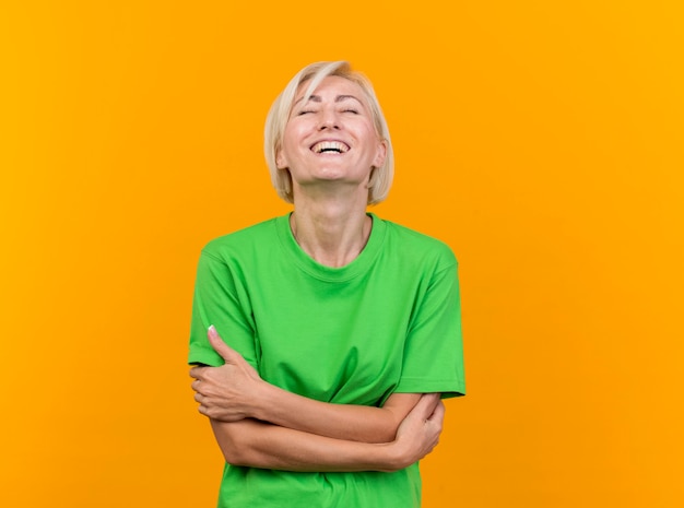 Joyeuse femme slave blonde d'âge moyen debout avec une posture fermée et riant les yeux fermés isolé sur fond jaune avec espace de copie