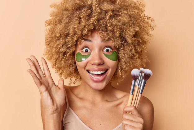 Joyeuse femme surprise aux cheveux bouclés garde la paume levée fixe les yeux sur écoute applique des patchs verts pour rafraîchir la peau tient des pinceaux cosmétiques isolés sur fond marron Concept de soins du visage