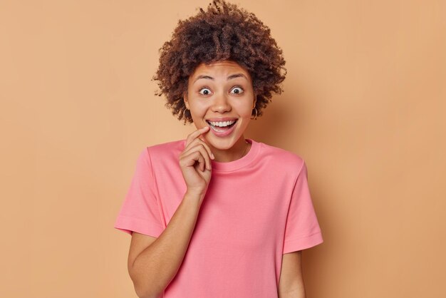 Joyeuse femme surprise sourit largement garde le doigt près de la bouche se sent étonné vêtu d'un t-shirt rose décontracté entend quelque chose de génial se surprend isolé sur fond marron. Émotions heureuses