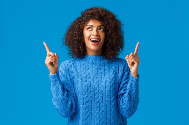 Joyeuse fille afro-américaine amusée célébrant avec des amis le nouvel an, joyeuses fêtes, regardant des feux d'artifice avec un sourire excité, pointant du doigt le regard étonné et joyeux, debout sur fond bleu.