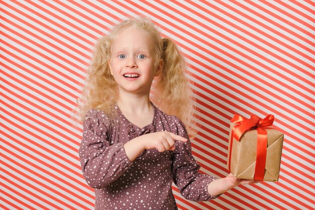Joyeuse fille d'âge préscolaire avec un cadeau sur fond rayé rouge, un cadeau avec un ruban rouge dans les mains d'une fille aux cheveux blonds, le lendemain de noël, des cadeaux pour l'anniversaire et noël, des cadeaux de vacances