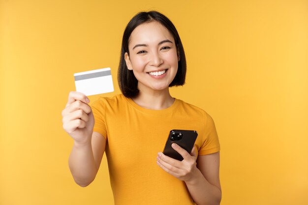 Joyeuse fille asiatique souriante montrant une carte de crédit et un smartphone recommandant des services bancaires par téléphone mobile debout sur fond jaune