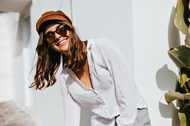 Joyeuse fille aux cheveux courts dans des lunettes brunes et une casquette se plie et sourit sur fond de bâtiment blanc et d'usine