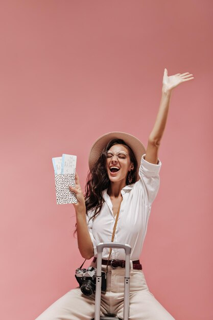 Joyeuse fille aux cheveux ondulés en chemise élégante pantalon beige avec ceinture et chapeau de paille se réjouit et tenant des billets sur fond rose