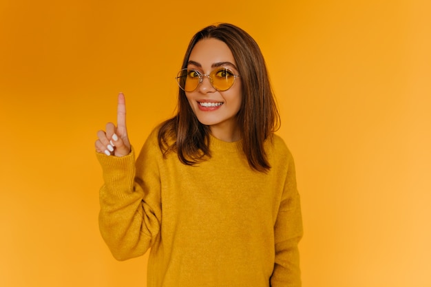 Joyeuse fille bronzée dans des verres posant sur un mur orange. Jolie jeune femme en pull tricoté.