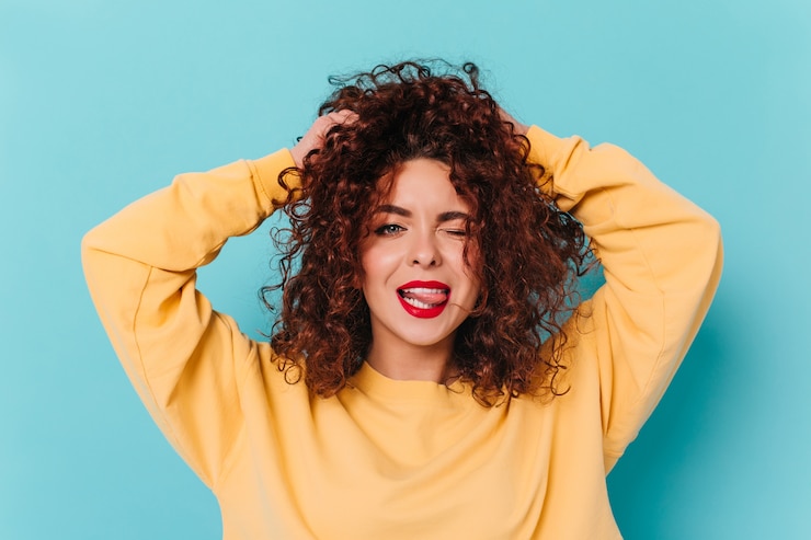 Photo gratuit joyeuse fille brune bouclée aux yeux bleus et rouge à lèvres clignote, touche les cheveux et montre la langue sur l'espace bleu.