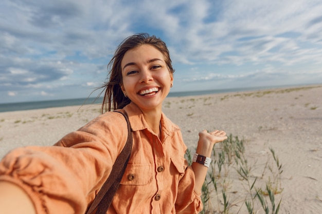 Joyeuse fille brune faisant autoportrait et profitant de vacances près de l'océan. Vacances, ambiance tropicale, chaudes journées d'été.