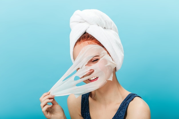 Joyeuse fille caucasienne décollant le masque facial. Photo de Studio de jeune femme insouciante faisant des soins de la peau sur fond bleu.