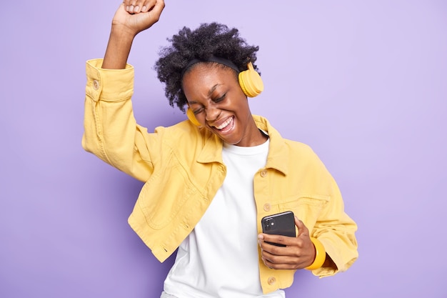 Joyeuse fille du millénaire aux cheveux bouclés danse insouciante aime playlsit préféré détient un téléphone mobile et des écouteurs modernes