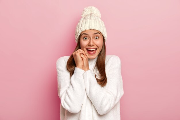 Joyeuse fille du millénaire aux cheveux noirs a une réaction heureuse aux bonnes nouvelles, sourit largement, porte un chapeau d'hiver chaud et un pull blanc confortable, a un regard enthousiaste, isolé sur un mur rose