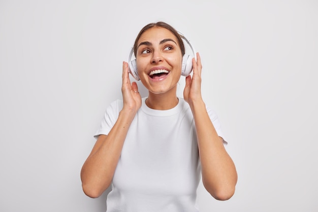 Joyeuse fille du millénaire écoute la piste audio préférée dans des écouteurs sans fil sourit largement a une expression heureuse habillée avec désinvolture isolée sur un mur blanc