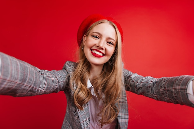 Joyeuse fille française aux cheveux longs prenant selfie. Dame blonde de bonne humeur en béret isolé sur un mur rouge.