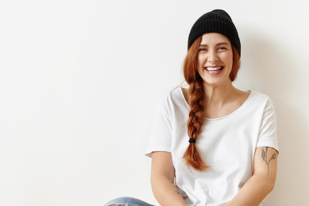 Joyeuse fille hipster mignonne insouciante avec tatouage assis sur le sol au mur blanc