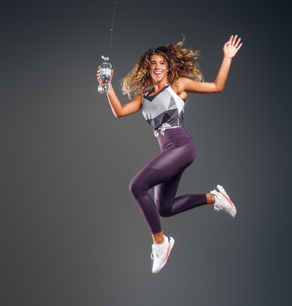 Joyeuse fille joyeuse saute avec une bouteille d'eau faisant des éclaboussures au studio photo sur fond sombre.