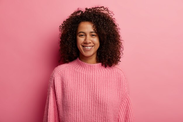 Joyeuse fille millénaire aux cheveux afro touffus sourit à pleines dents, profite d'un moment heureux, regarde avec joie la caméra, porte un pull tricoté, isolé sur fond rose.