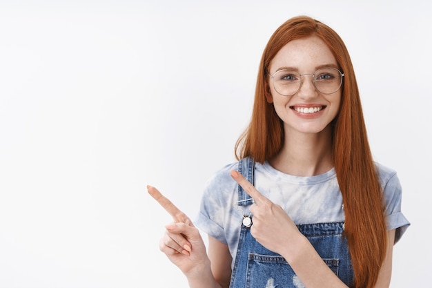 Joyeuse fille rousse sincère et amicale aux yeux bleus, taches de rousseur, longs cheveux roux pointant vers la gauche, souriante polie et sociable présente la promo, stand mur blanc amusé aide joyeuse et heureuse