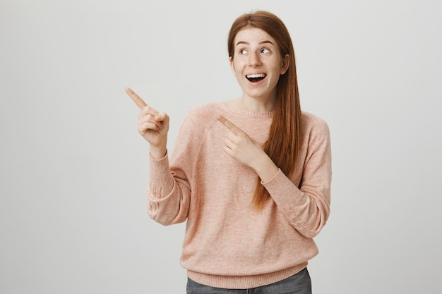 Joyeuse fille rousse souriante pointant les doigts dans le coin supérieur gauche