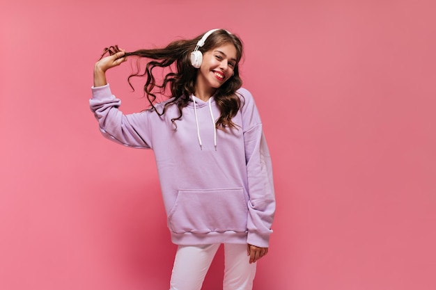 Joyeuse fille en sweat à capuche violet et pantalon blanc sourit sincèrement Bonne femme bouclée écoute de la musique dans les écouteurs et pose sur fond rose isolé