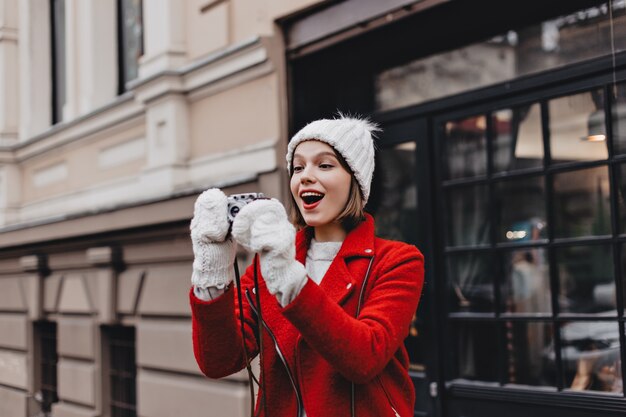 Joyeuse fille en veste rouge, bonnet tricoté et mitaines prend une photo de la ville avec un appareil photo rétro.