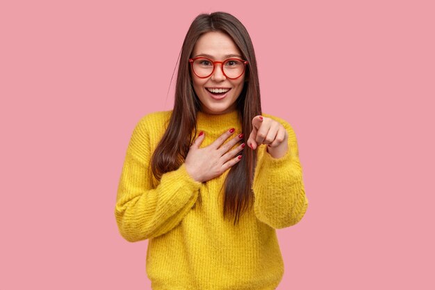 Joyeuse heureuse jeune femme métisse pointe directement à la caméra, rit avec de bonnes émotions, garde la main sur la poitrine, ne peut pas arrêter les sentiments