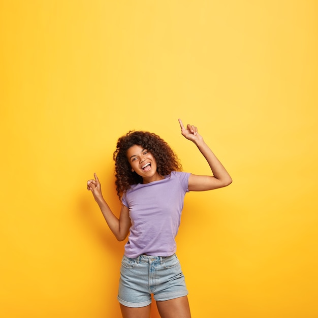 Joyeuse heureuse jolie femme à la peau sombre avec des cheveux crépus, lève les mains sur une bonne musique vibrée, a une silhouette mince