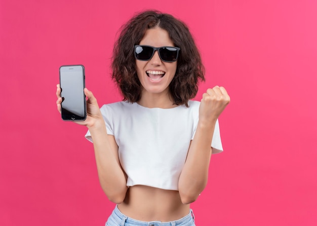 Joyeuse jeune belle femme portant des lunettes de soleil et tenant un téléphone mobile sur un mur rose isolé