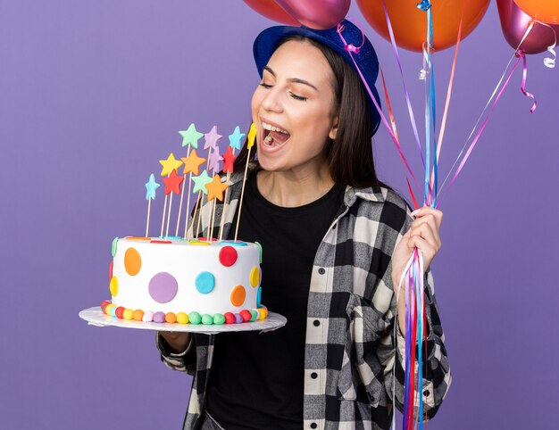Joyeuse jeune belle fille portant un chapeau de fête tenant des ballons avec un gâteau isolé sur un mur bleu