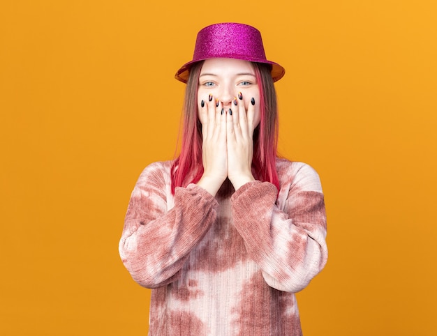 Joyeuse jeune belle fille portant un chapeau de fête visage couvert de mains isolées sur un mur orange