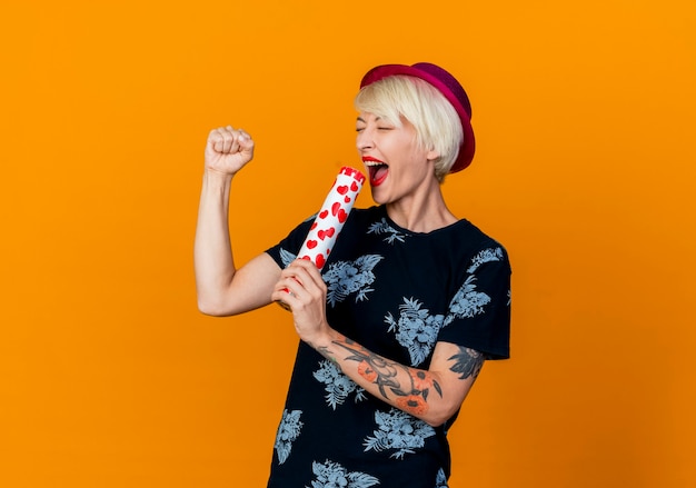 Joyeuse jeune blonde party girl wearing party hat chant à l'aide de canon à confettis comme microphone avec les yeux fermés isolé sur fond orange avec copie espace