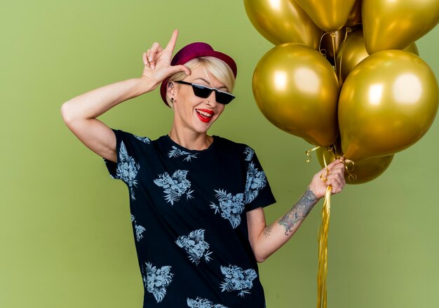 Joyeuse jeune blonde party woman wearing party hat et lunettes de soleil tenant des ballons faisant le geste perdant isolé sur mur vert olive