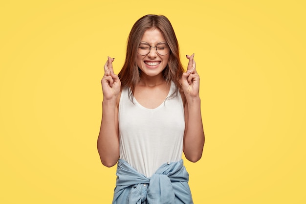 Joyeuse jeune brune avec des lunettes posant contre le mur jaune