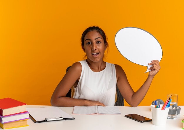 Joyeuse jeune écolière assise au bureau avec des outils scolaires tenant une bulle de discussion