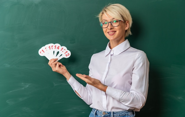 Joyeuse jeune enseignante blonde portant des lunettes en classe debout devant le tableau montrant et pointant vers le nombre de fans regardant la caméra