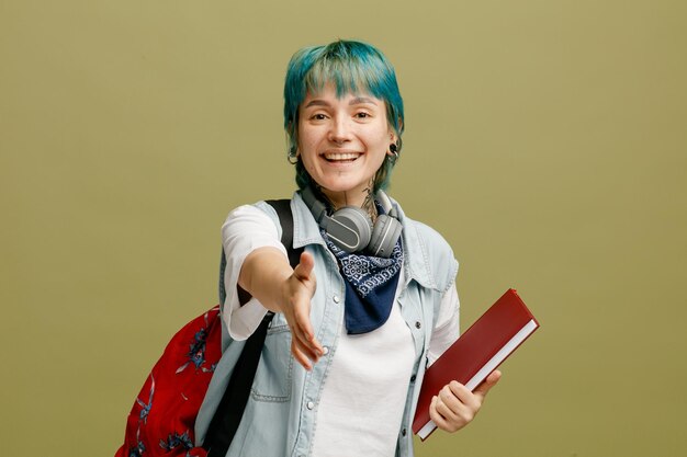 Joyeuse jeune étudiante portant des écouteurs et un bandana sur le cou et le sac à dos tenant un carnet de notes regardant la caméra montrant un geste agréable de vous rencontrer isolé sur fond vert olive