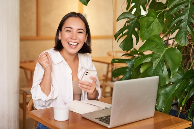 Joyeuse jeune femme asiatique gagnante en célébrant la victoire ou la réussite recevoir de bonnes nouvelles sur son téléphone portable