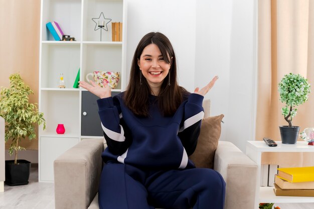 Joyeuse jeune femme assez caucasienne assise sur un fauteuil dans le salon conçu à la recherche et montrant les mains vides