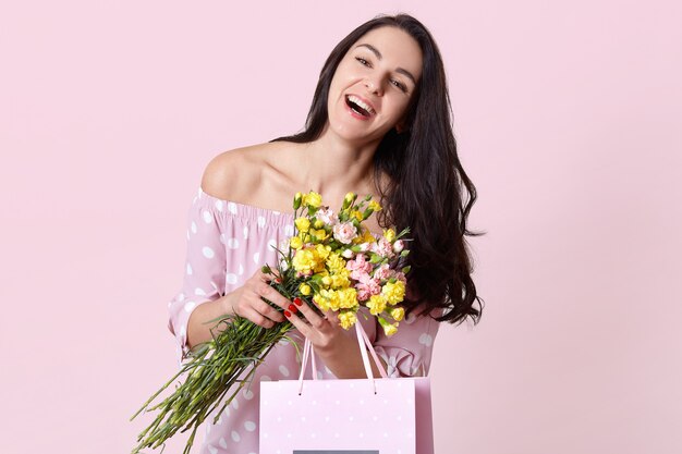 Joyeuse jeune femme aux cheveux noirs sourit joyeusement, vêtue de vêtements élégants, heureuse de recevoir un cadeau pour l'anniversaire, détient un bouquet de fleurs, des modèles sur rose. Concept de temps de printemps