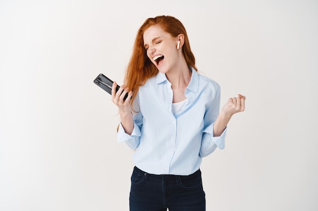 Joyeuse jeune femme aux longs cheveux roux chantant et écoutant de la musique dans des écouteurs sans fil, s'amusant, tenant un smartphone, debout sur un mur blanc