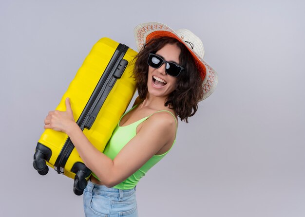 Joyeuse jeune femme belle voyageur portant chapeau et lunettes de soleil et tenant la valise sur un mur blanc isolé avec espace copie