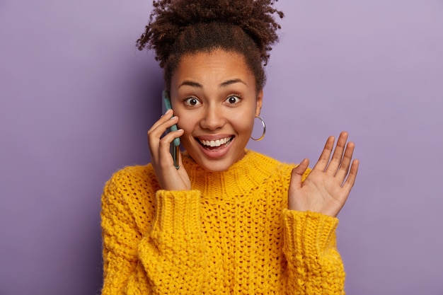 Photo gratuite joyeuse jeune femme bouclée parle au téléphone, heureuse d'entendre de bonnes nouvelles, des gestes pendant la conversation, soulève la paume, porte des boucles d'oreilles et un pull jaune, aime parler décontracté, isolé sur fond violet