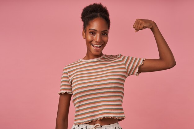 Photo gratuite joyeuse jeune femme brune attrayante avec une coiffure chignon décontractée en levant la main tout en démontrant son fort biceps, debout sur un mur rose