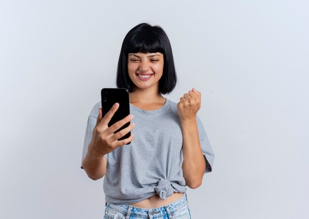 Joyeuse jeune femme caucasienne brune garde le poing et tient le téléphone