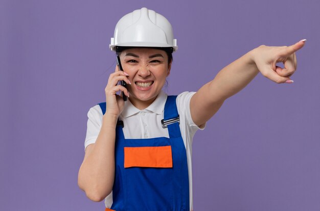 Joyeuse jeune femme de construction asiatique avec un casque de sécurité blanc parlant au téléphone et pointant sur le côté